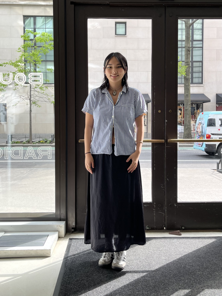 Student wears a blue and white striped short sleeve button up shirt, long black slip skirt, and white and black Adidas Samba sneakers while standing in the lobby of the University Center.
