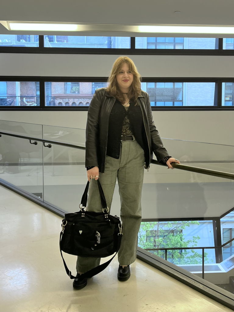 Student wears a black leather jacket, black cardigan, green pants, and holds a large black bag while leaning on a glass railing.