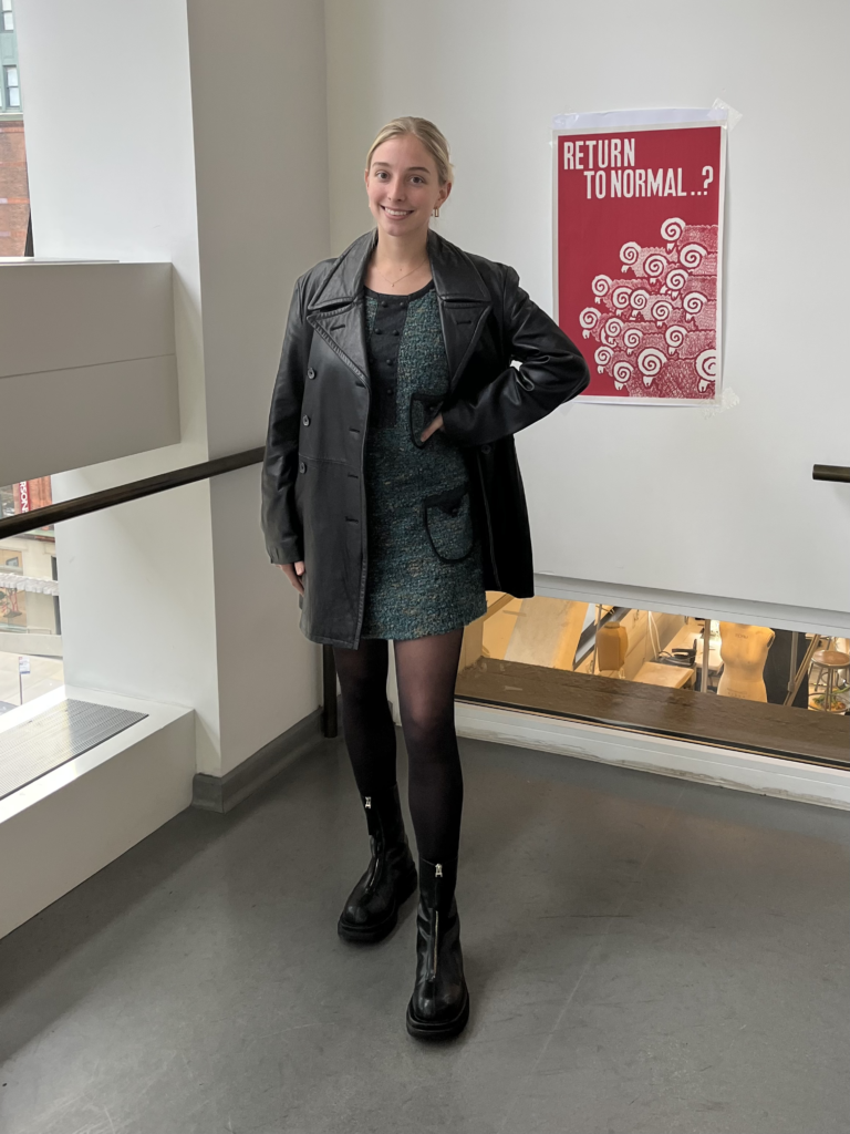 Student wears a black leather jacket, dark green tweed dress, black mesh stockings, and black boots while standing with her hand on her hip.