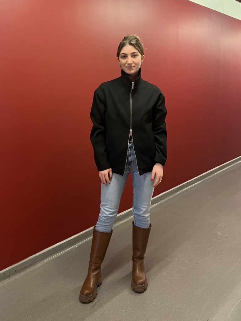 Student wears a black double zip sweater, blue skinny jeans, and tall brown boots while standing in front of a red wall.