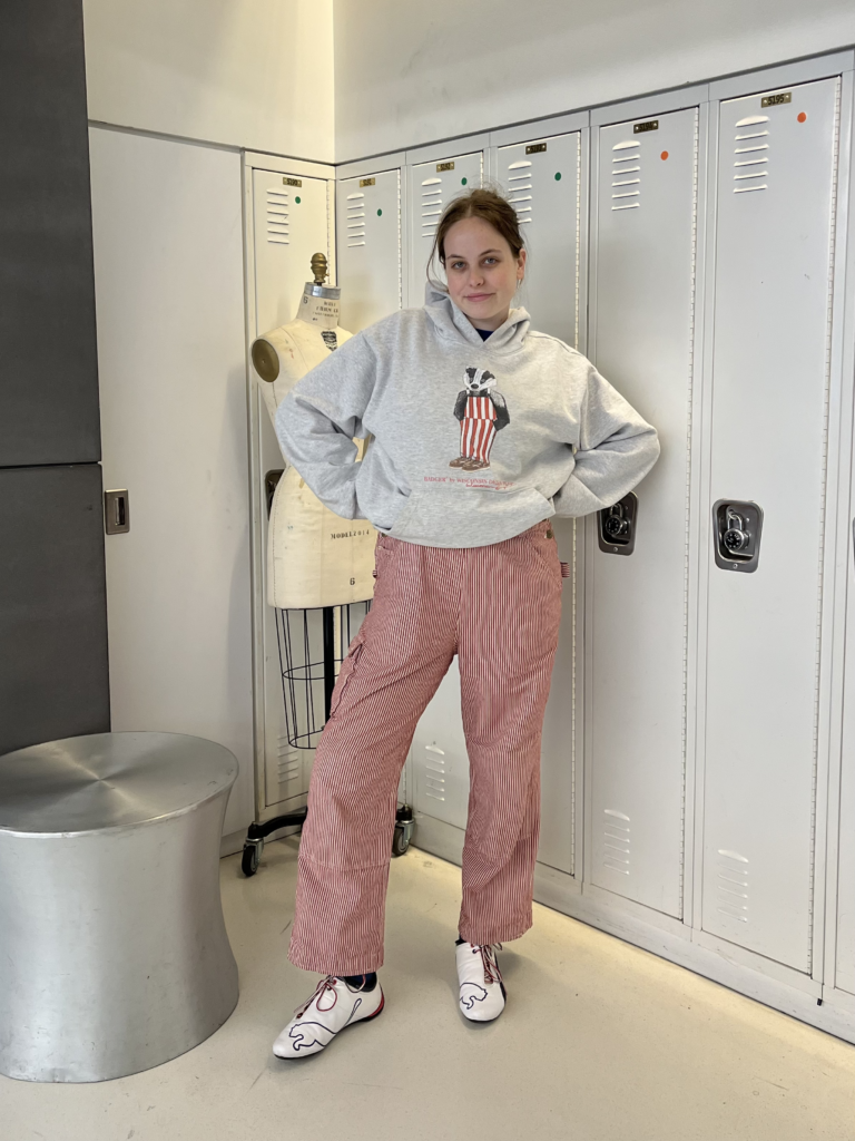 Student wears a gray hoodie with a badger graphic over red and white striped overalls and white Puma sneakers while standing in front of white lockers.