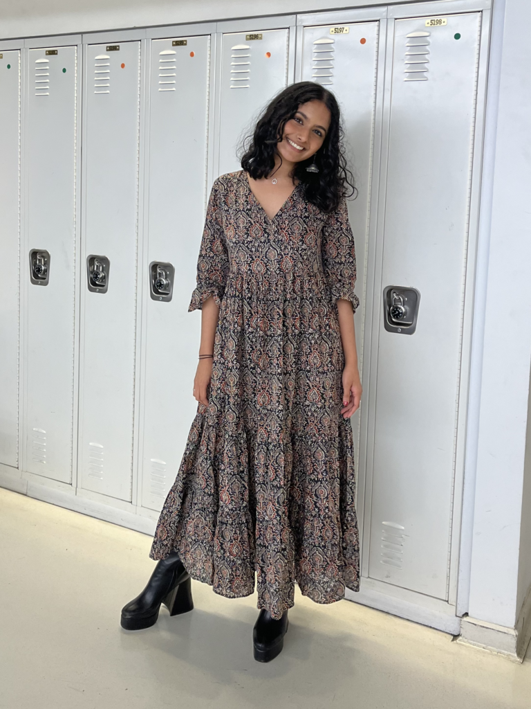 Student has long dark hair, a long flowy patterned brown dress, and black platform boots while standing in front of white lockers.