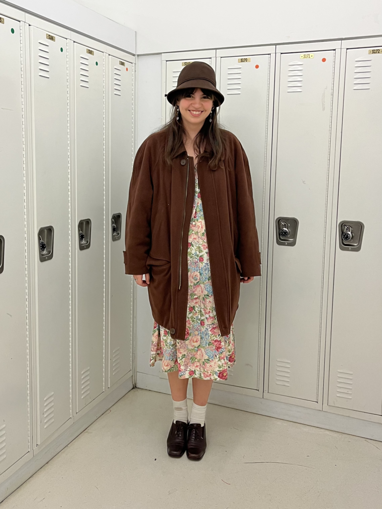 Student wears a floral dress, long brown coat, and brown cloche hat while standing in front of white lockers.