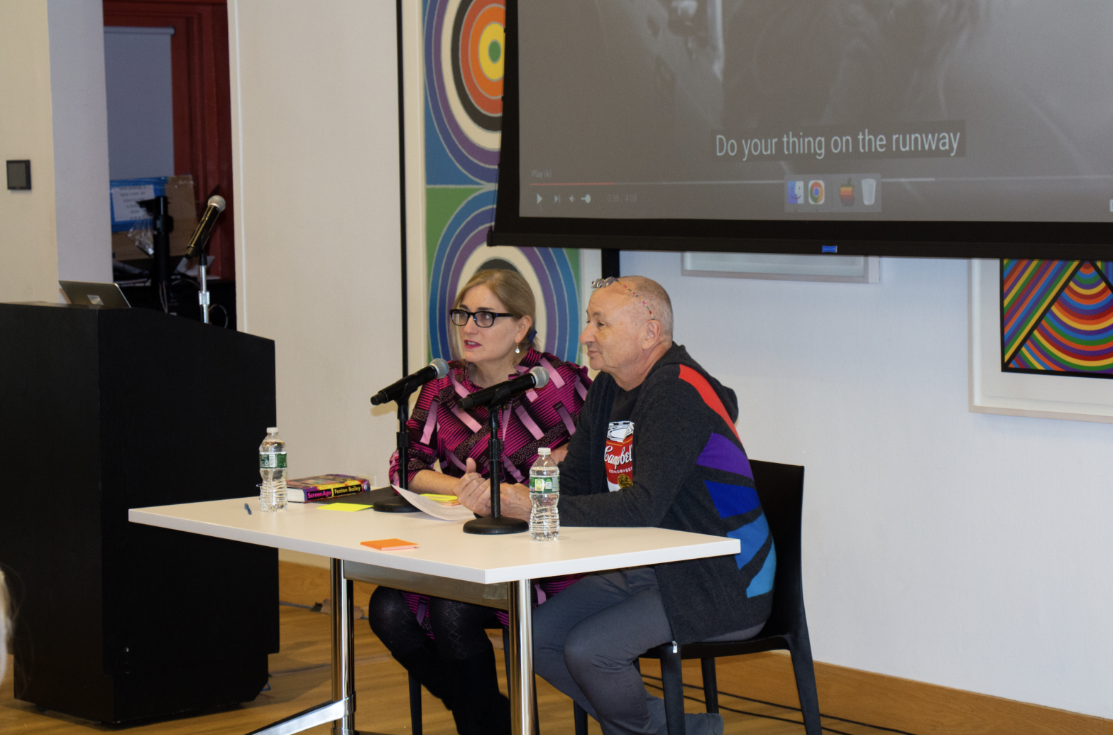 Fenton Bailey and Liesel Schinger sitting in front of an audience, with their notes strewn across a white table top. In the background RuPaul’s music video Supermodel is projected.