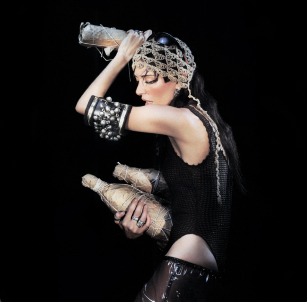 Alt text: Artist Caroline Polachek stands alone against a pitch black backdrop with the left side of her body facing the camera. She wears an ornate headpiece and a large, embellished bangle on her arm. Holding three bottles wrapped in paper, one held slightly above her head, she looks down with a dramatic expression on her face.
