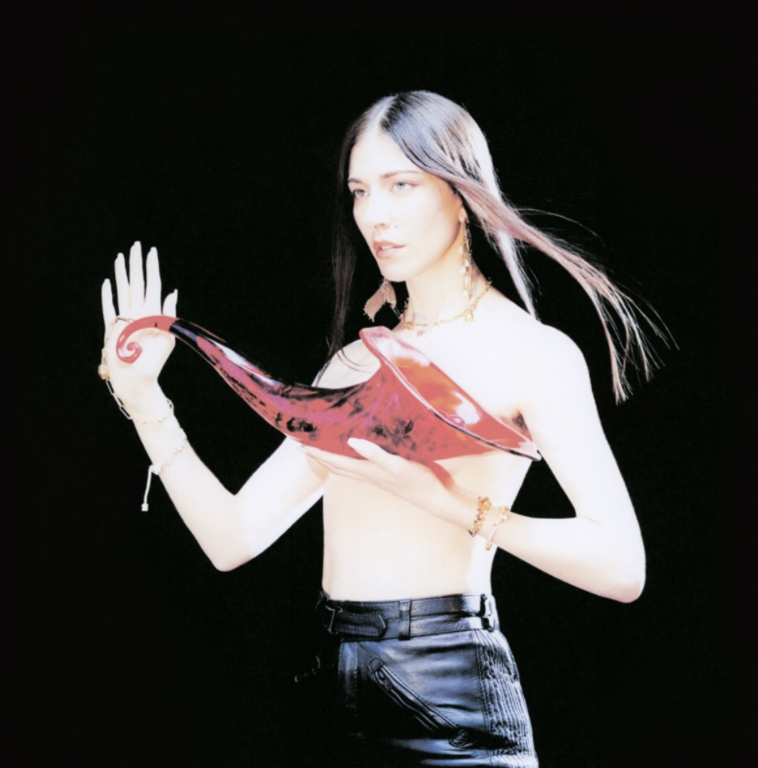 Artist Caroline Polachek stands alone against a pitch black backdrop, holding an ornate glass instrument.