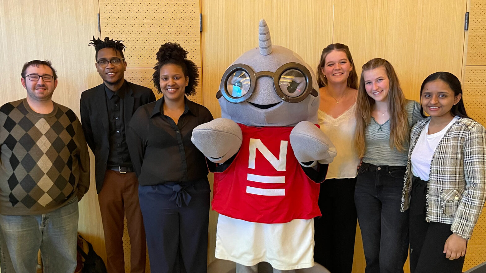 A photograph of The New School’s mascot, Gnarls, with five Student Senators and a USS advisor standing next to them. There are three people on each side of Gnarls. All are standing in front of a brown wall in a New School building.