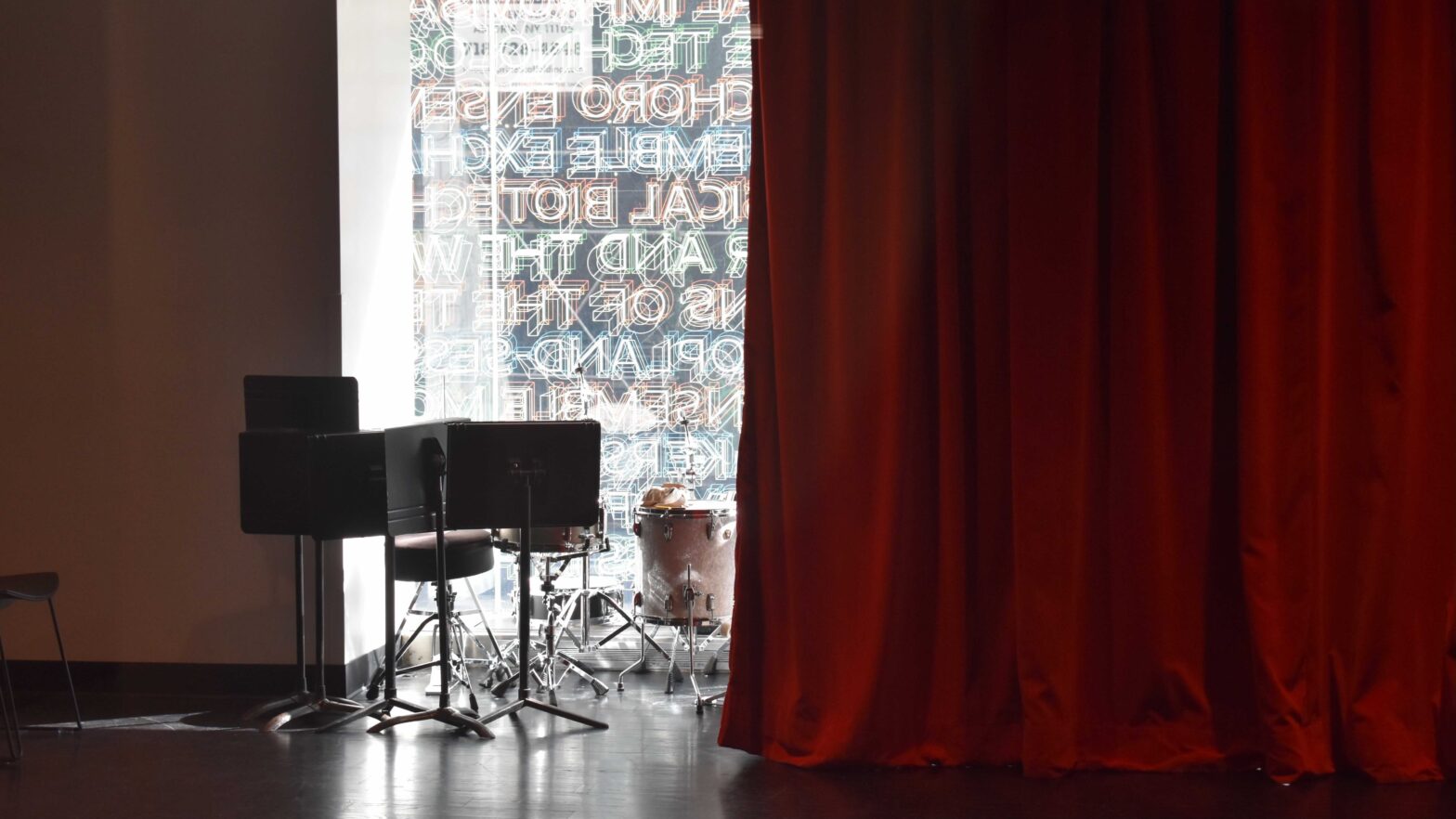 A photograph of a dimly lit room with four black music stands and a drum set in front of a window with colorful print on it. Half of the window is covered by a dark red velvet stage curtain.