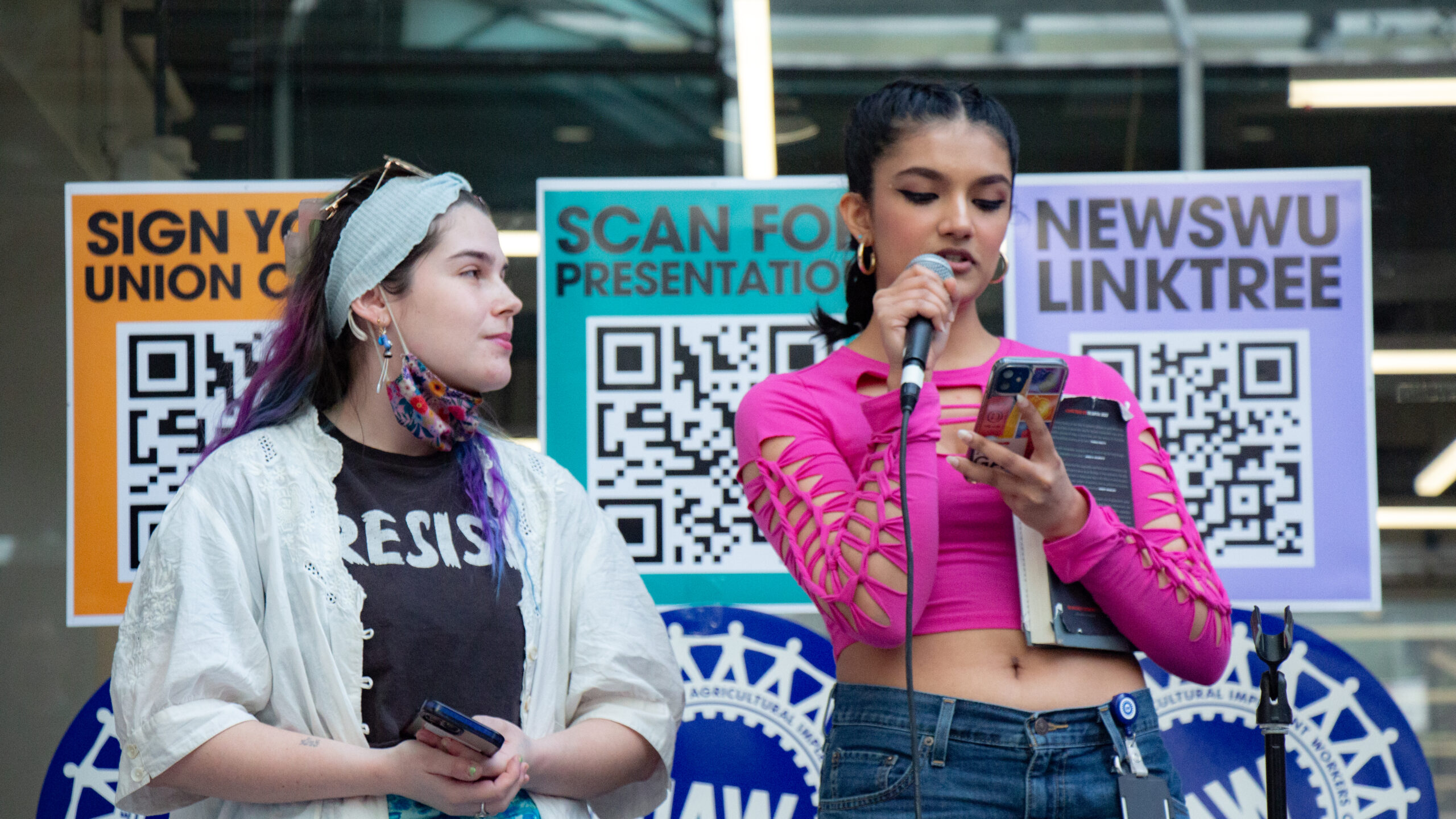 Two people, the one on the right holding a microphone, stand in front of three large, brightly colored signs with QR codes hung to a glass building wall. The first sign reads “Sign Your Union Card.” The second sign reads “Scan for Presentation.” The third sign reads “NewSWU Linktree.”