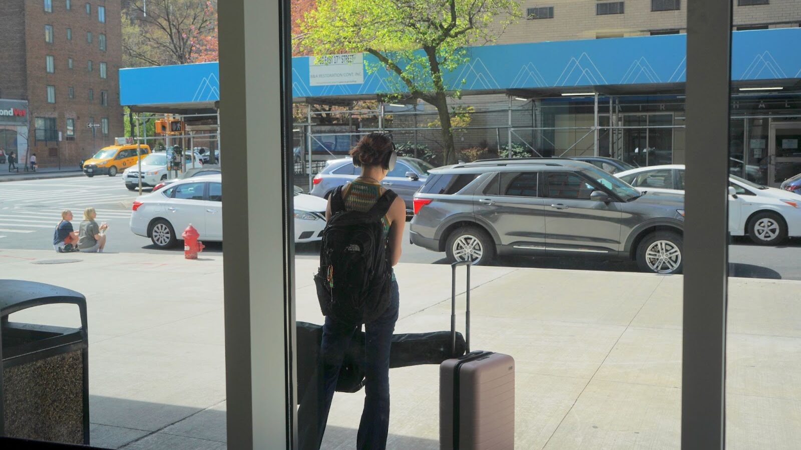Student, seen through a window, waits for a taxi on street with luggage on left and a guitar case in hand.