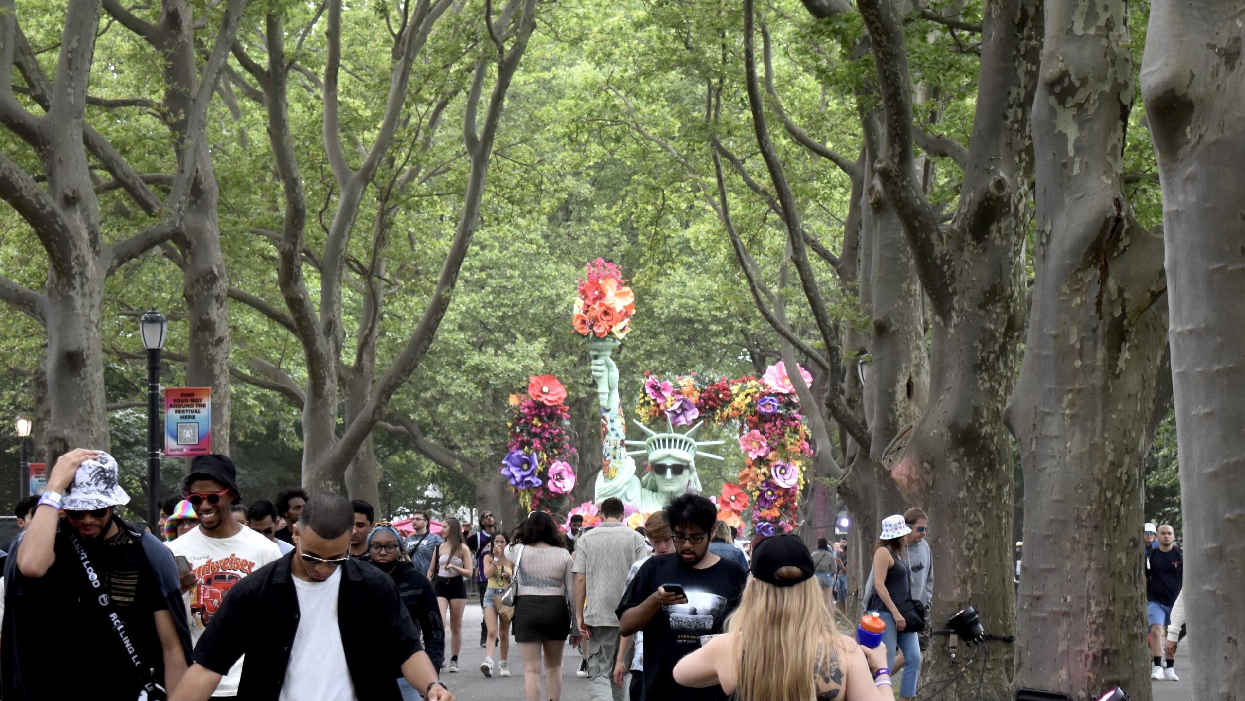 Concert goers walk around a sculpture of the statue of liberty wearing sunglasses, holding a torch of flowers, and surrounded by a flower arch.