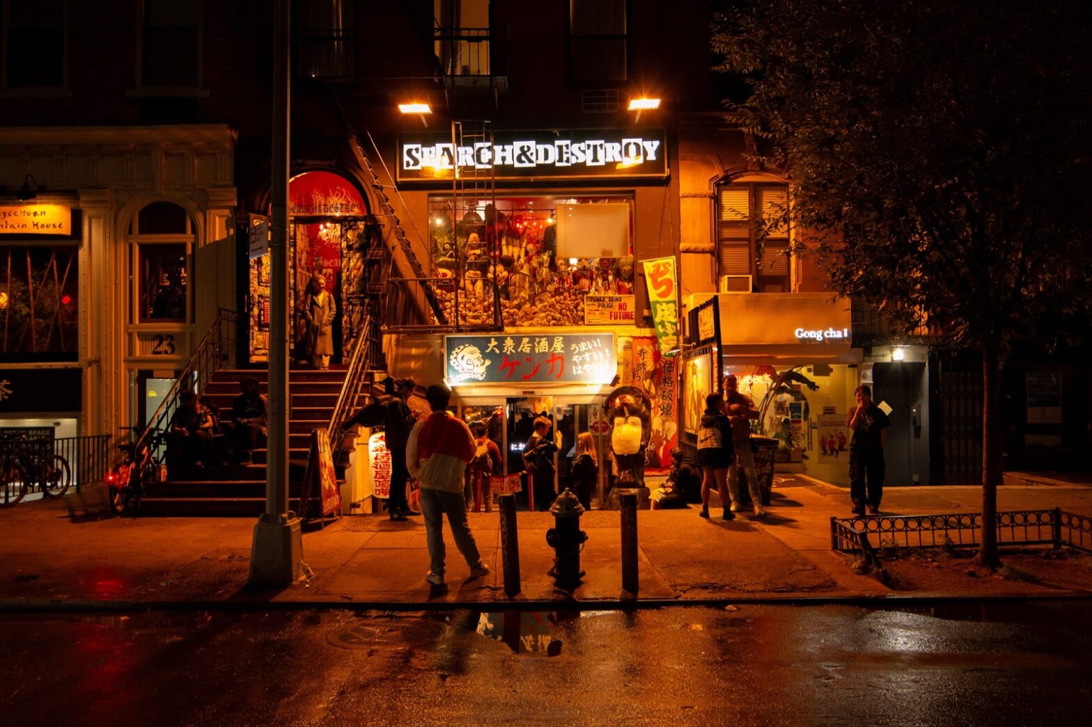 The entryway to the restaurant KENKA taken from the street at night. The restaurant’s entrance is below street level underneath a storefront which reads “Search and Destroy” The entryway emits warm light from lanterns and lit signs with Japanese text. People stand on the sidewalk in front of the doors surrounding the establishment.