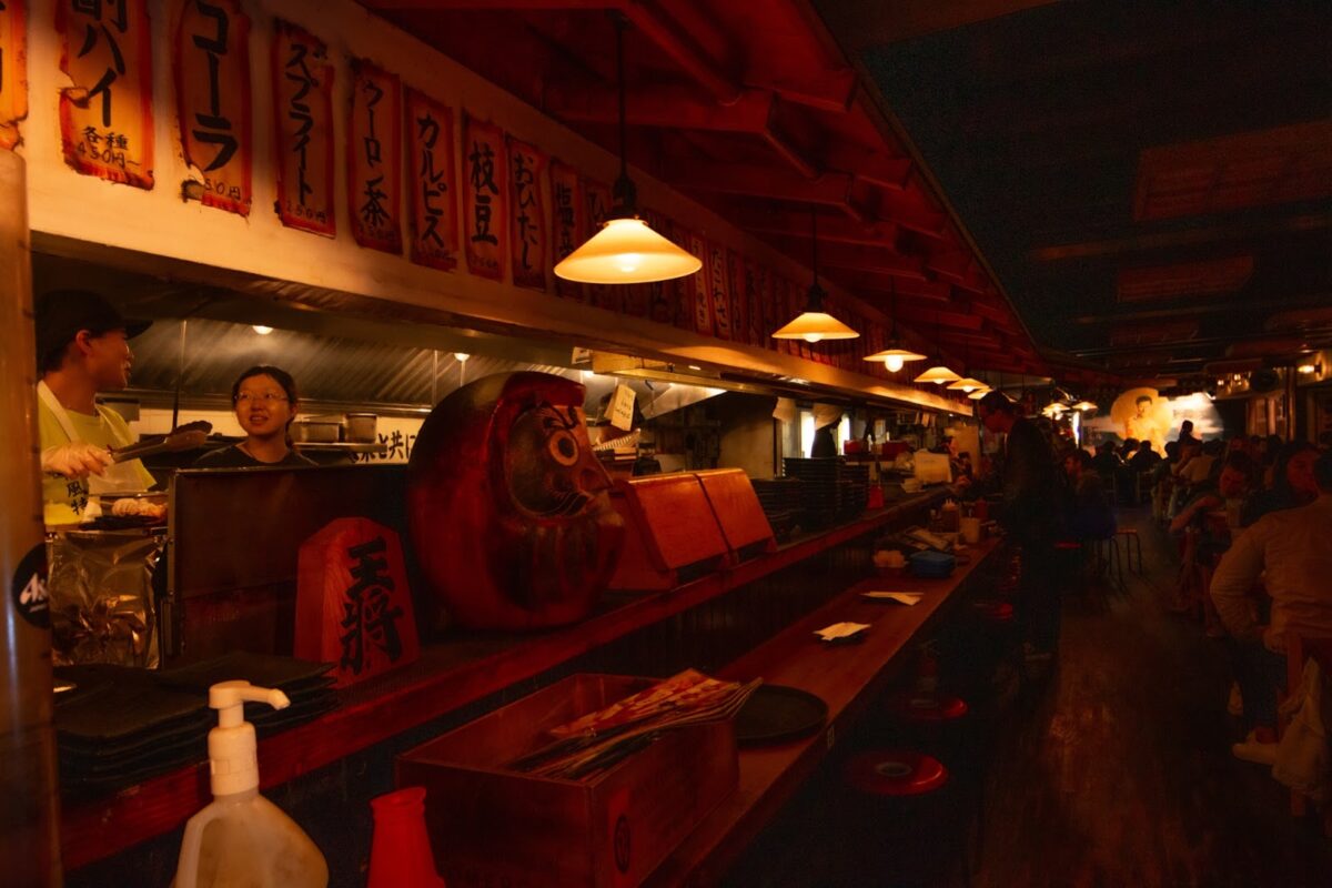 Inside the restaurant KENKA, a long wooden bar with seating separates the main dining room from the kitchen. The light is low and the wall above the bar is fully covered in ripped posters with Japanese lettering. Sitting on the bar is a Japanese doll head called a Daruma. In the far distance at the back of the room a mural of a man is partially visible. 

