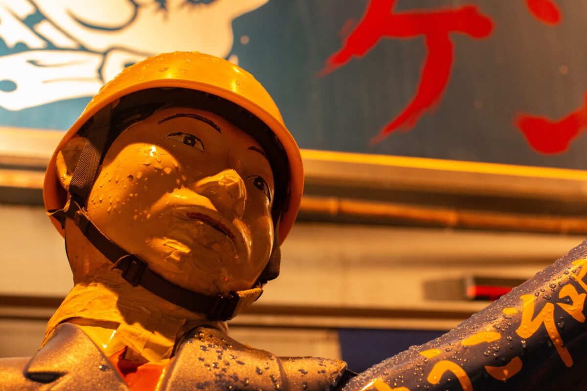 A life size vintage crossing guard statue wearing a yellow helmet stands in front of the door to the restaurant. Only the face and arm are visible from the close angle. 