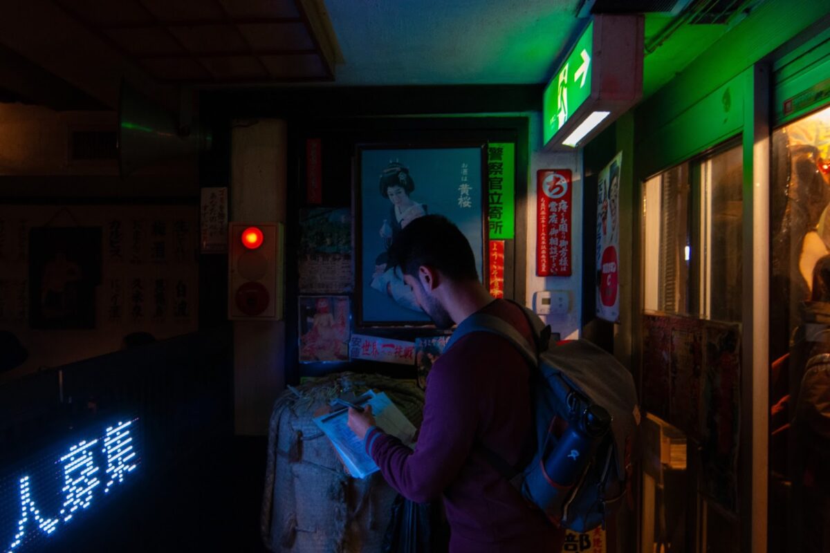 A green exit sign illuminates the waiting area of the restaurant. There are posters and led signs decorating the walls. A man wearing a backpack stands inside writing  his name on a clipboard.
