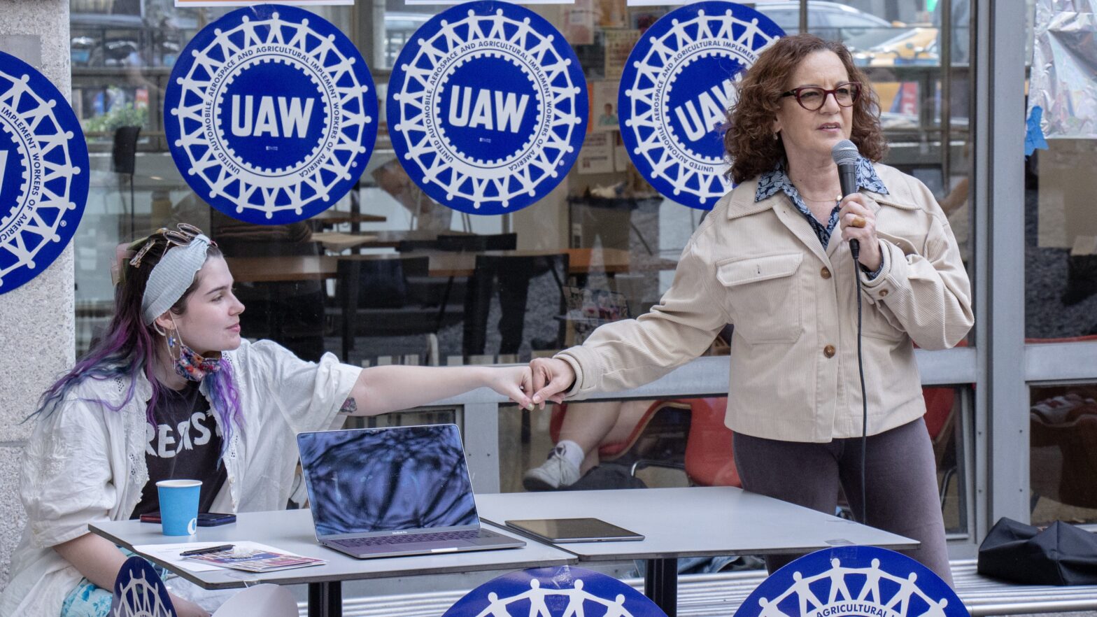 Alt Text: Behind a table, two people fist bump. One is sitting and is wearing a white jacket and bandana. The other is standing and speaking into a microphone. Behind them are four blue and white UAW union signs and orange, teal, and purple signs with QR codes.