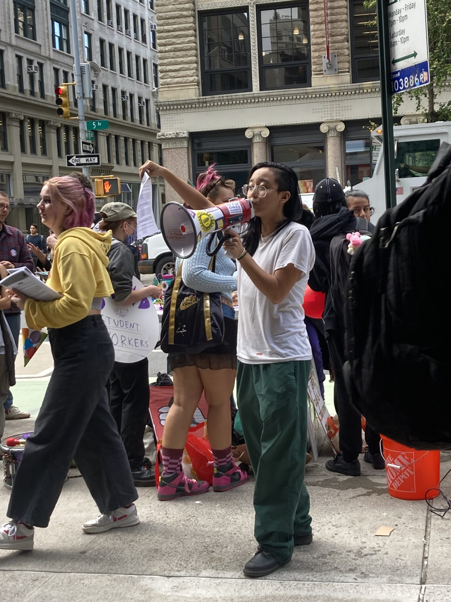 A person in green pants and a white shirt is speaking into a megaphone and holding a sheet of paper. A small crowd of roughly six people stands behind them, talking and holding various signs and bags.