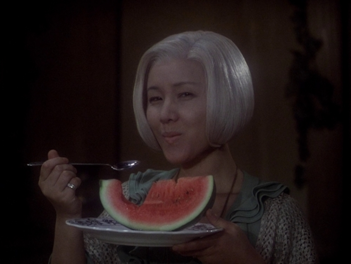 A screen capture from “House”(1977) shows a woman with gray hair eating a slice of watermelon.
