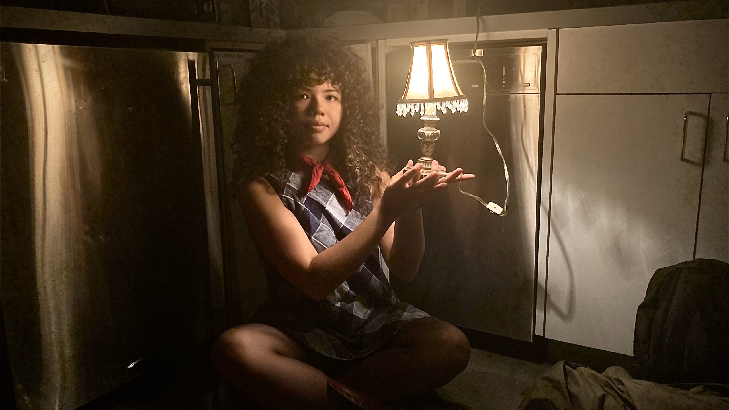 Image of Lamp Club founder Jackie McVorran sits criss cross on the floor of the Lang pantry, leaning against a dishwasher and a cabinet door. She is sporting a red necktie and blue geometric patterned dress, and she gazes into the camera, illuminated by the glow of the miniature lamp she’s holding.