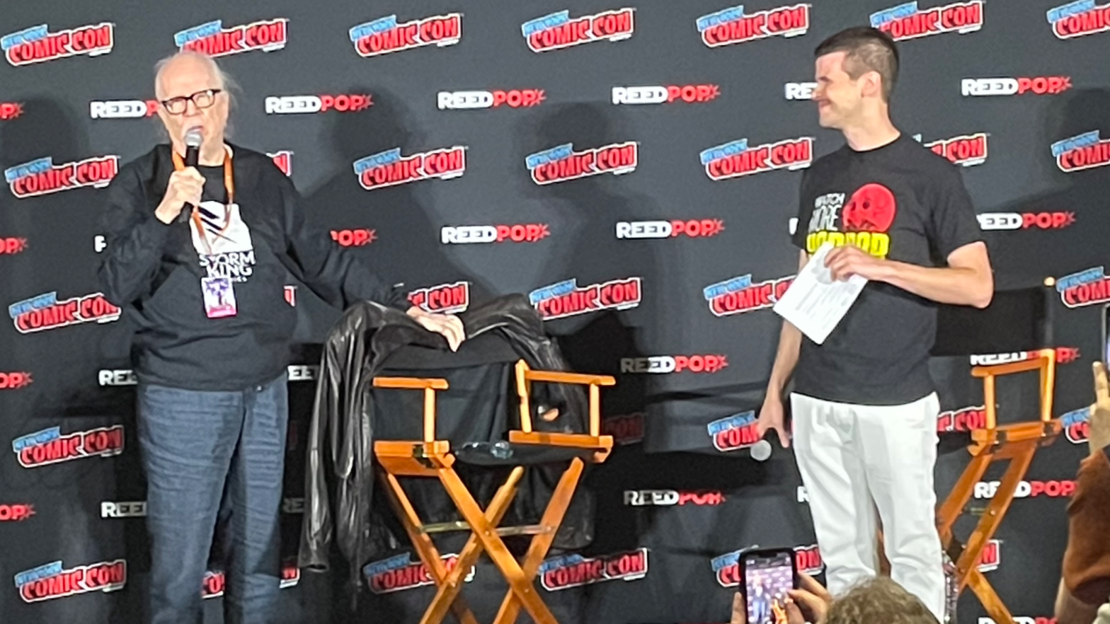 John Carpenter and his moderator standing on a stage inside the Javits Center in New York City holding microphones with a chair in between them.