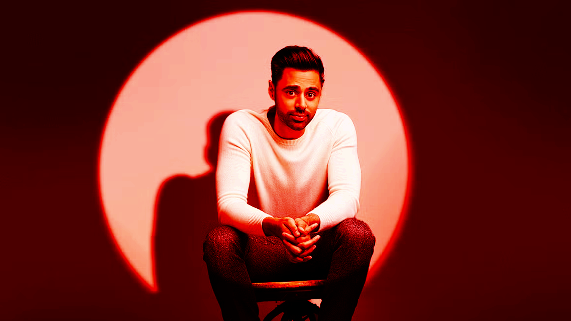 Hasan Minhaj sits in front of a bright red circular light