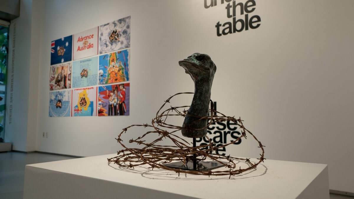 Sculpture of a duck head emerging from a spool of rusted barbed wire. Bright colored grid of tea towels displayed out of focus on the wall behind the sculpture. Photo by Avery Palmer