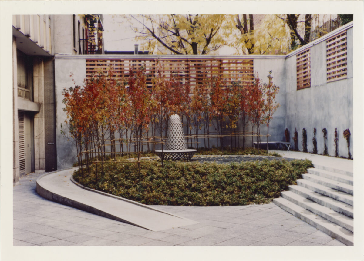 Trees with red leaves and green shrubbery centered around a metal sculpture in a courtyard.
