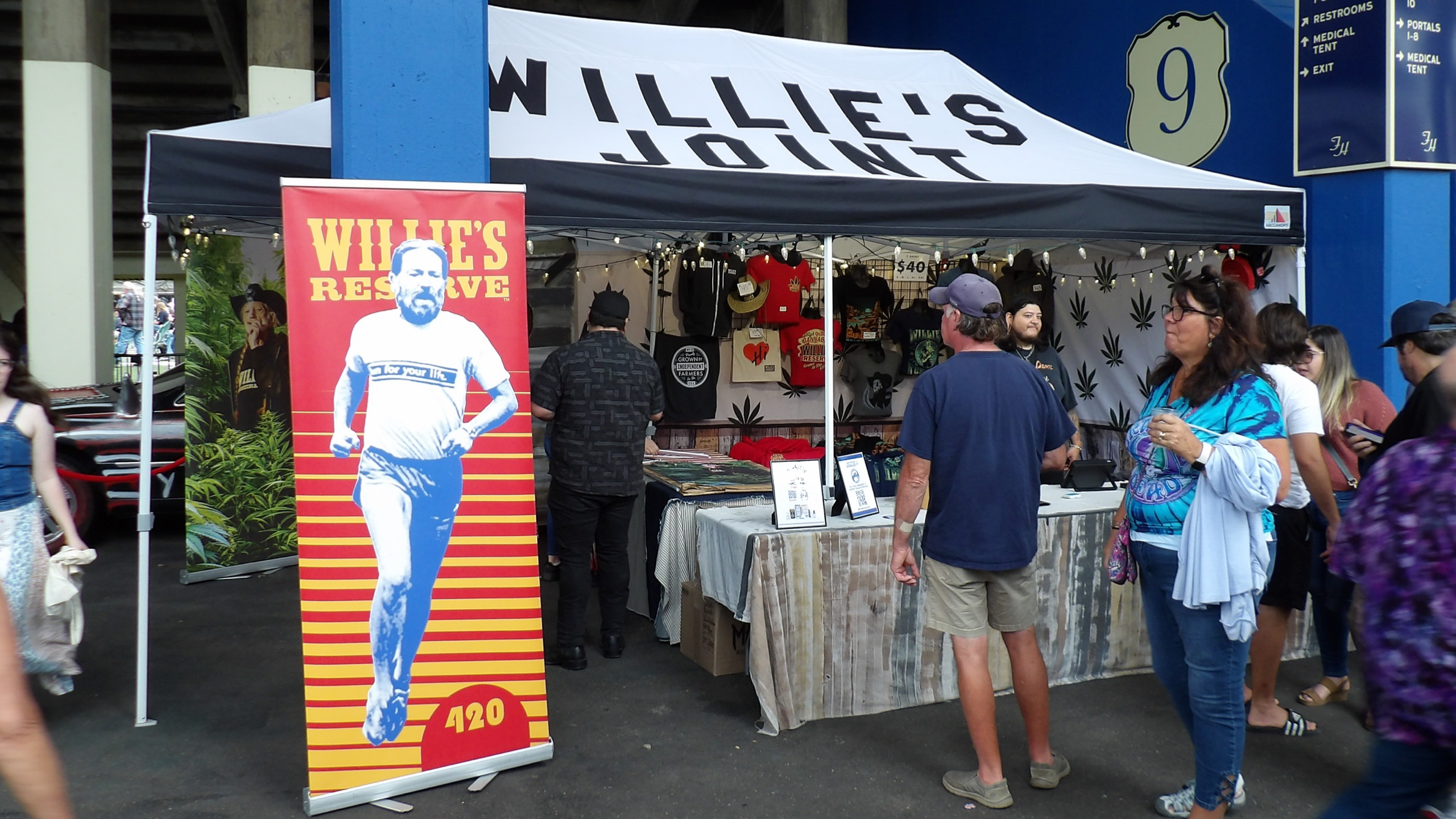 People outside of a merch tent, browsing Willie Nelson shirts next to a sign depicting a younger Willie Nelson running.