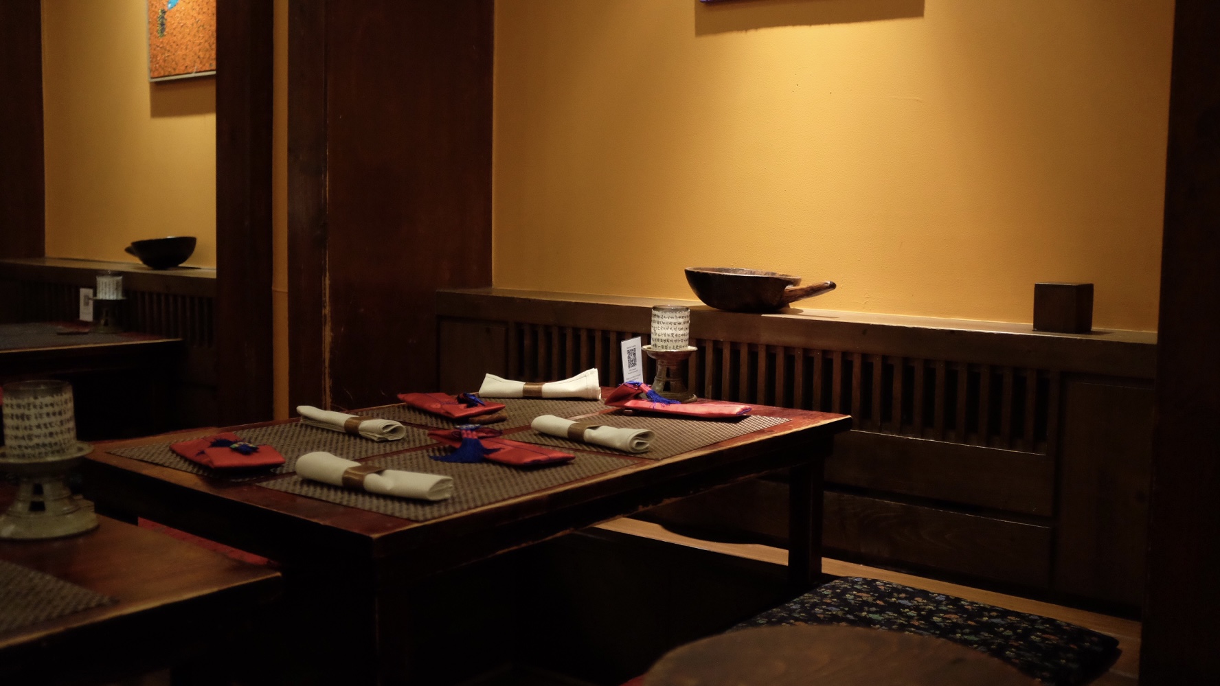 A photo of a table at Hangawi. The walls are painted a warm, deep yellow shade, and the wooden table is set with place settings and woven place mats, and surrounded by Korean artifacts. Small built-in benches have floral cushions on them for added padding and flare.