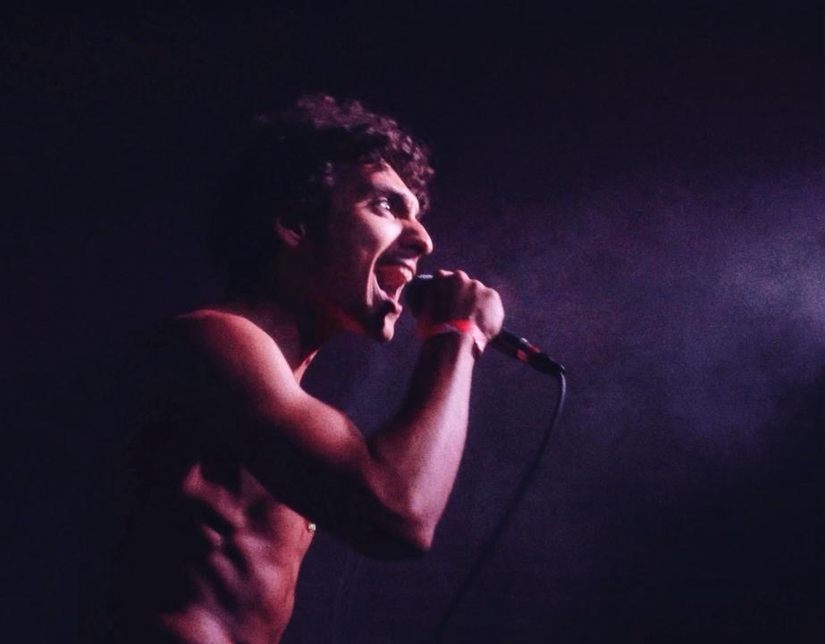 Nory Aronfield stands shirtless against a dark purple background, gripping a microphone while a spotlight shines on him as he sings.
