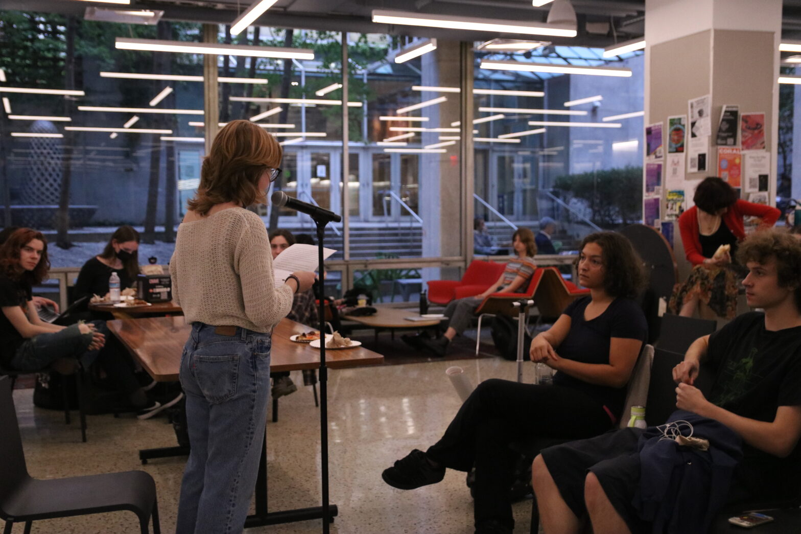 A student reads her writing in front of an student audience. She wears a white sweater and blue jeans.