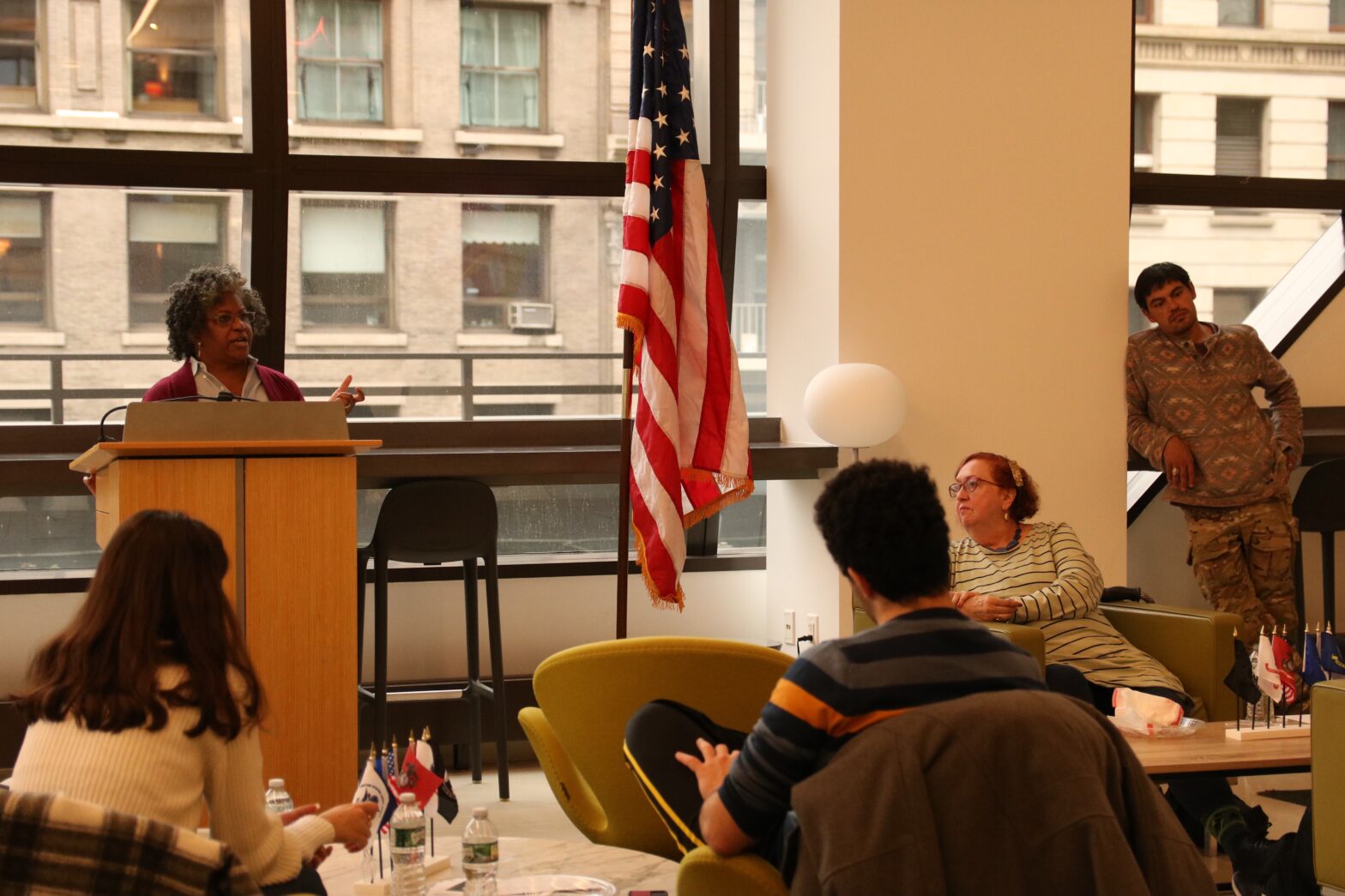 Woman in a purpl e sweater at a podium speaks to a small crowd next to a large American flag on a pole.