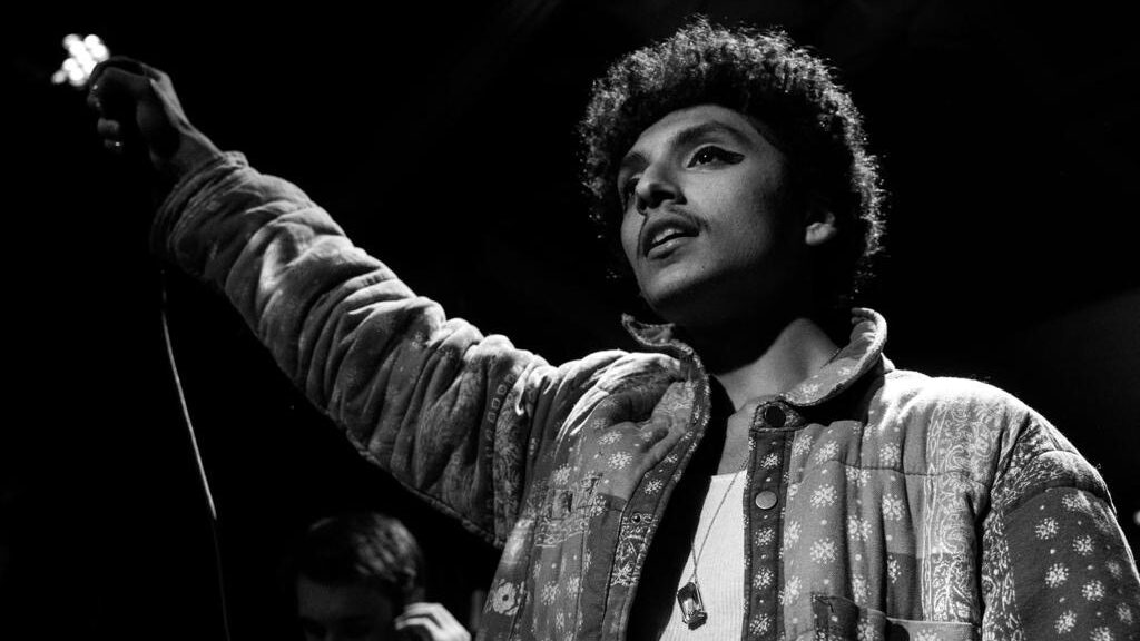 Black and white photo of Nory Aronfeld as he stands before the crowd with his right hand on the microphone.