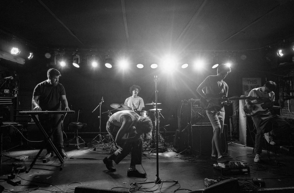 Black and White photo of Nory Aronfeld as he kneels towards the stage, the spotlights shining on an accompanying guitarist, drummer, bassist, and pianist who play behind him.