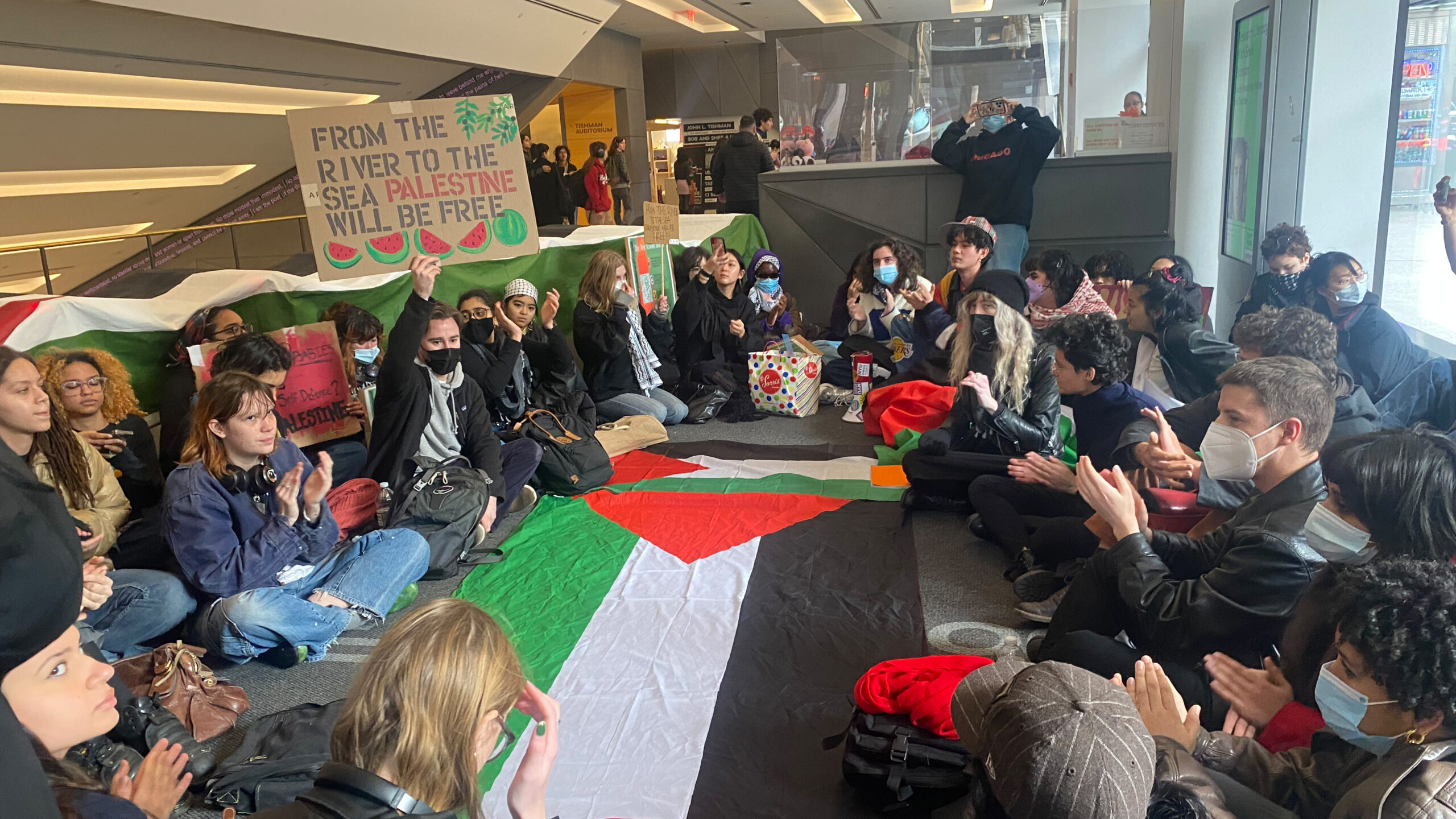 Students sit in a circle in front of the turnstiles at the University Center. In the center of the circle is one large Palestinian flag. One student to the left holds a sign that reads “From The River To The Sea, Palestine Will Be Free.”