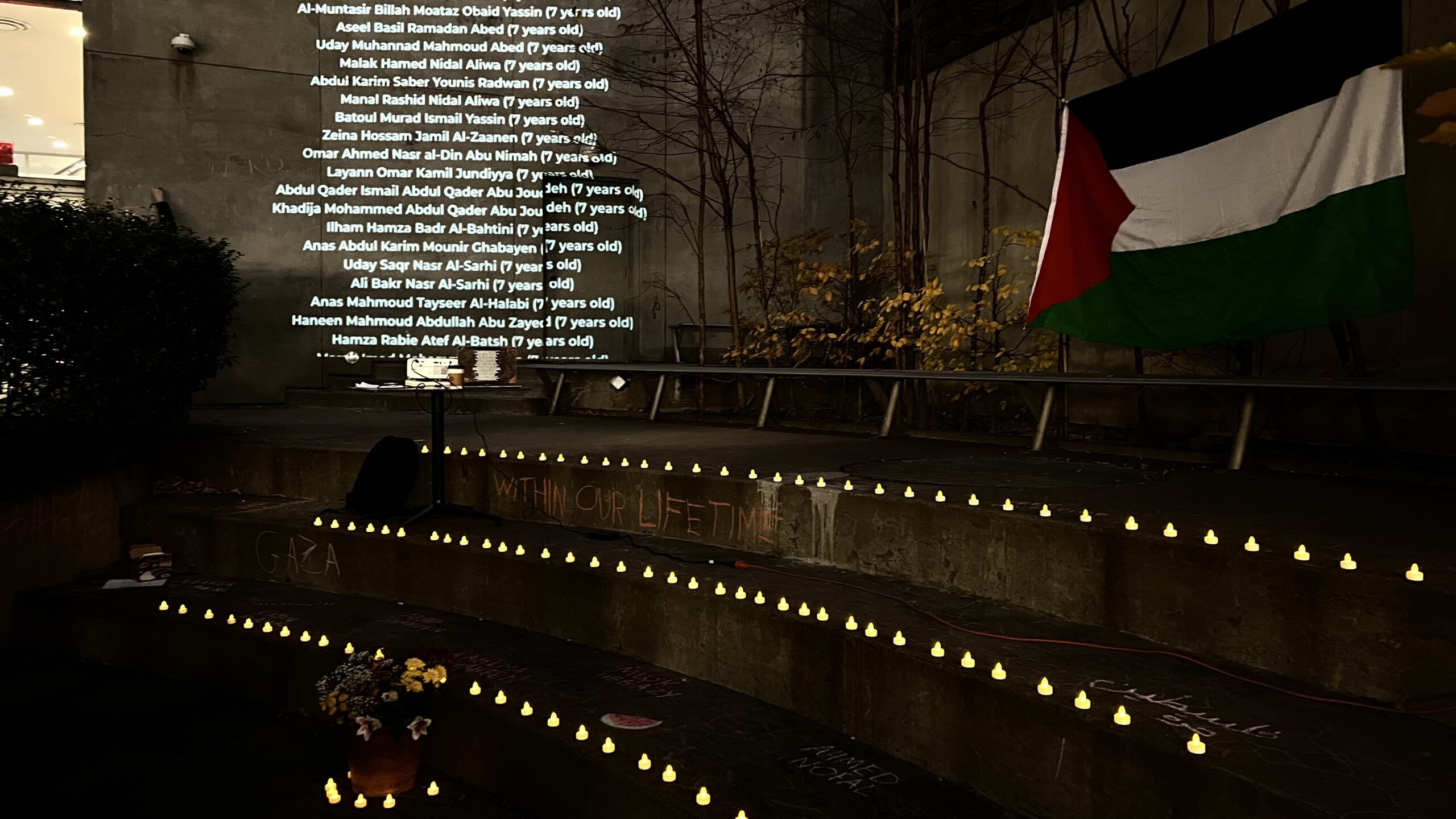 Steps in Lang Courtyard at night with candles and flowers on them and projected screen of names of the fallen to the left and Palestinian flag hung by the trees in the back