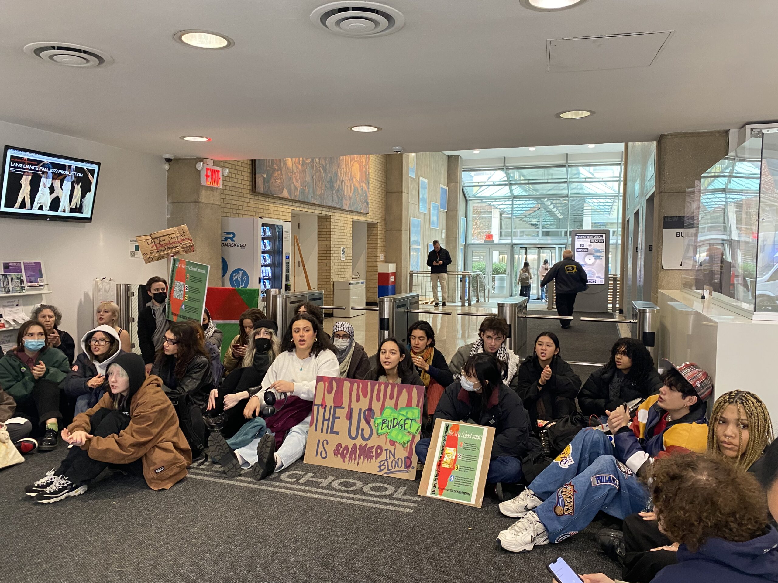 A group of demonstrators seated in front of Alvin Johnson/J.M. Kaplan Hall on East 12th Street. In the middle, a sign says “The US budget is soaked in blood.”