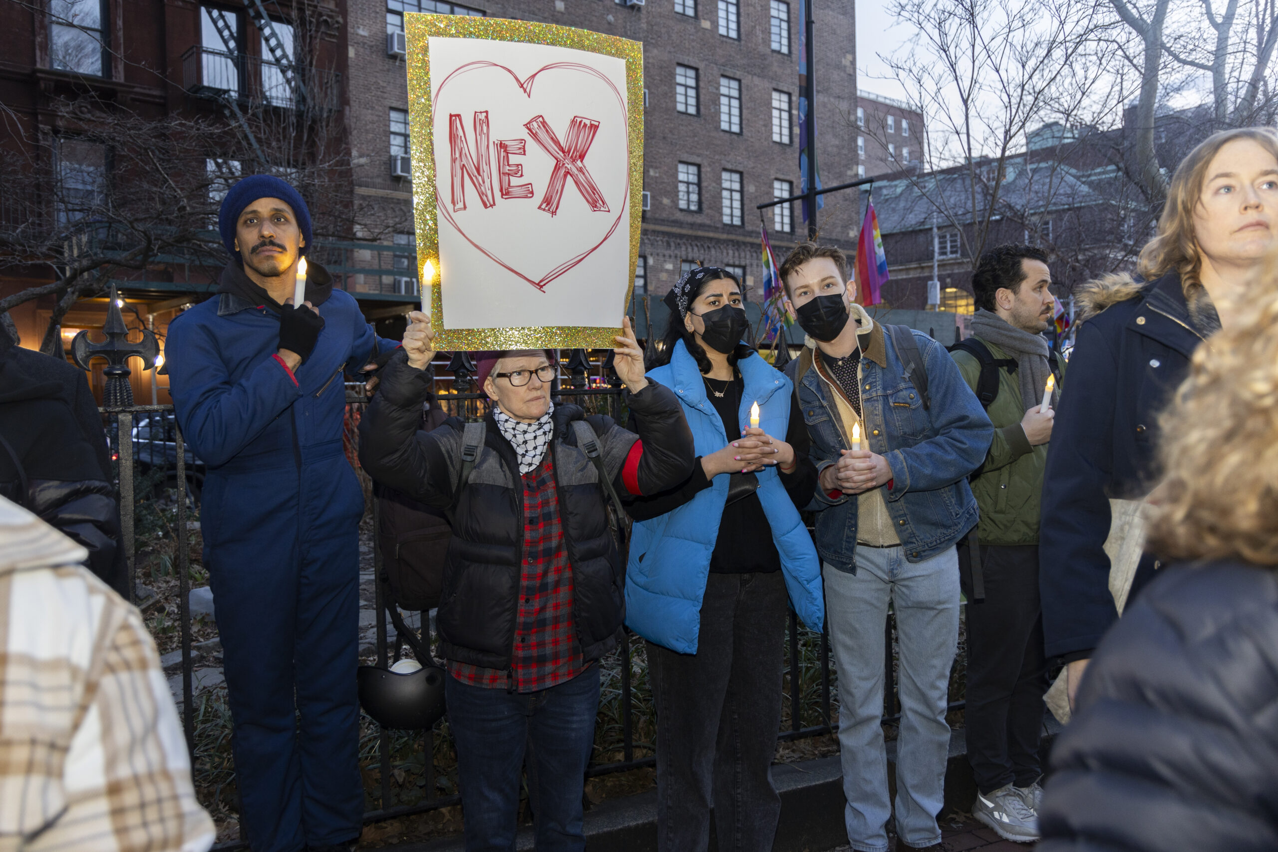 Person holds a sign that reads “NEX” with a heart drawn around it. Around them are others holding candles.