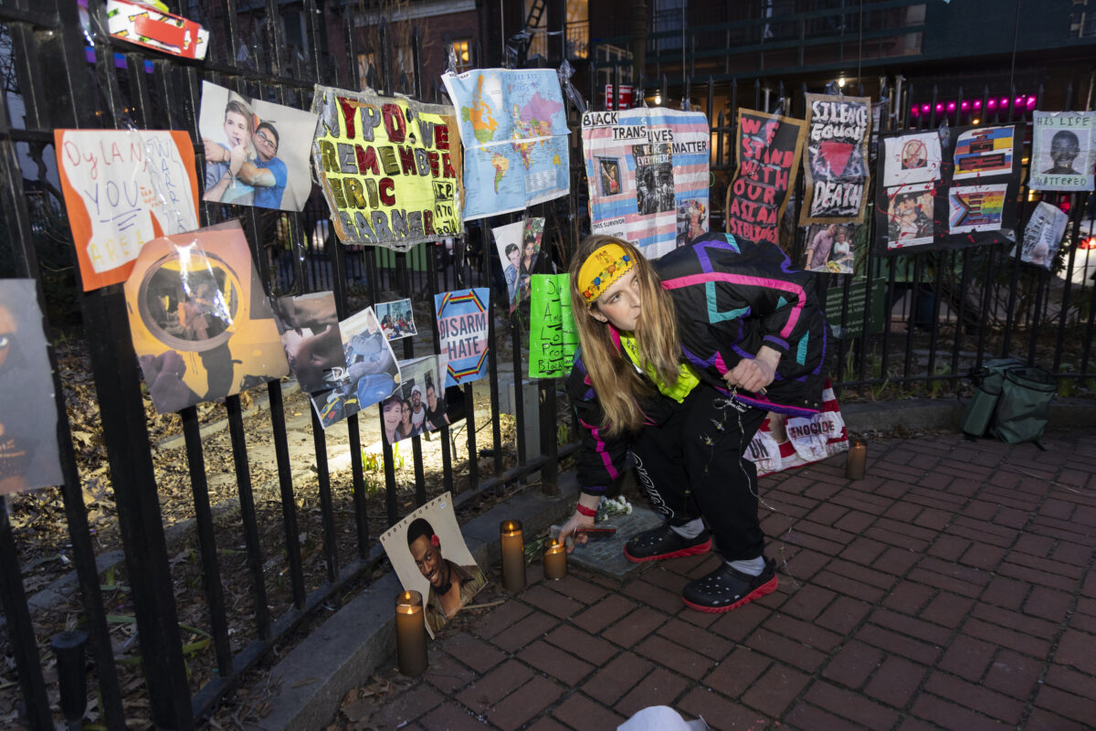 Image of a person kneeling down to place a candle. Behind them is a fence with memorial posters honoring queer people who have died.