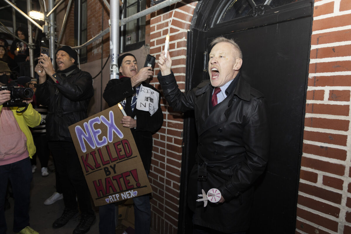 Image of a man yelling as he holds up a candle. Beside him, a man films him with his phone and holds a sign reading 'Nex killed by hate! RIP nex'
