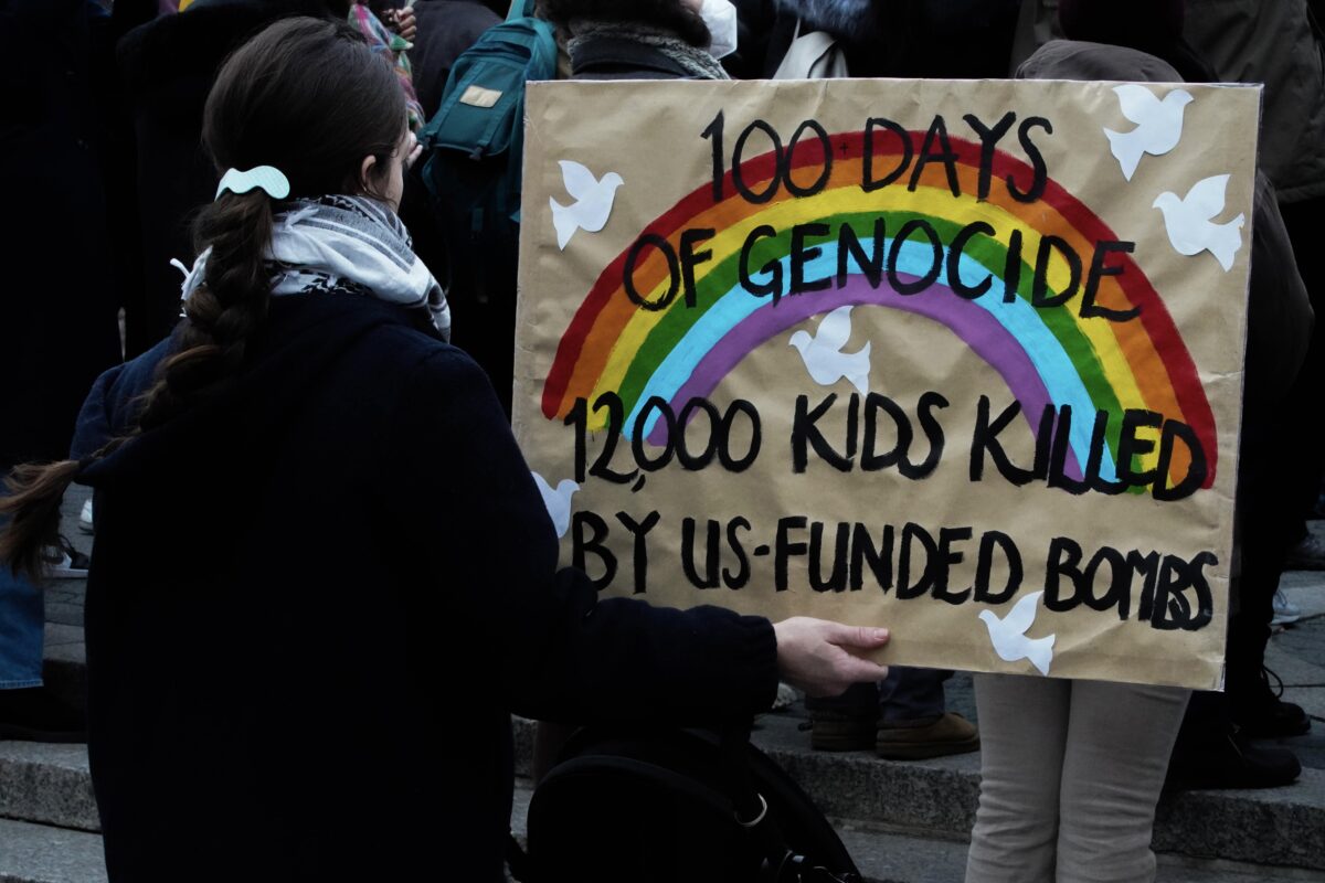 A woman with a braided ponytail wearing a keffiyeh and pushing a stroller and her child stands with her back facing the camera holding a large khaki sign decorated with white doves, a rainbow, and thin black letters reading “100 days on genocide 12,000 kids killed by us-funded bombs”