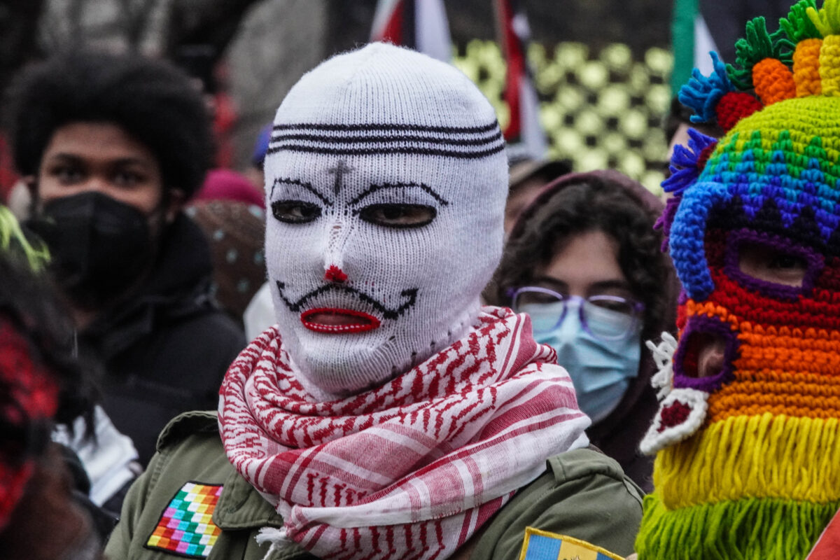 Protester wearing white knitted mask with a black cross, eyebrows,thin mustache and and around eyes with a red dot and nose and outline of mouth looks directly into the camera. Protester wearing an eccentric rainbow knitted mask stands to their left
