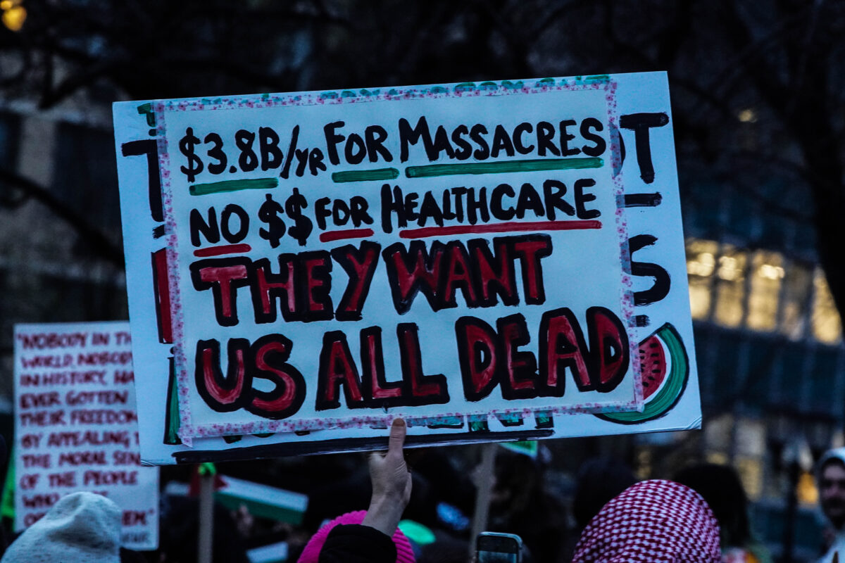 Sign reads “$3.8B/yr For Massacres No $$ For Healthcare with “they want us all dead” below in large bold red font