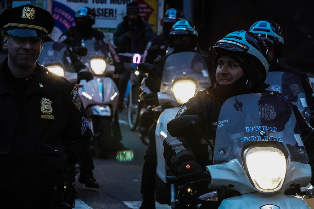 Five NYPD officers riding motorbikes with glowing headlights ride along side and an out of frame crowd of protesters the cop riding in front and an officer walking on foot to his right are smirking at each other 
