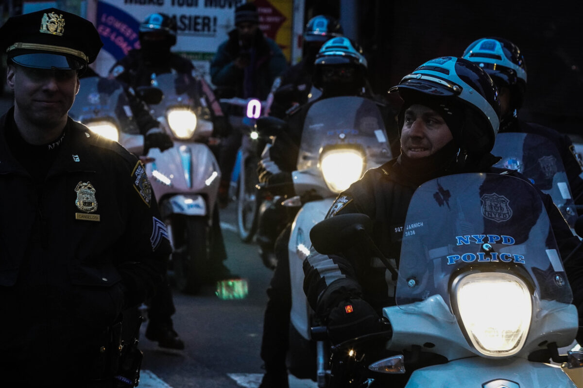 Five NYPD officers riding motorbikes with glowing headlights ride along side and an out of frame crowd of protesters the cop riding in front and an officer walking on foot to his right are smirking at each other