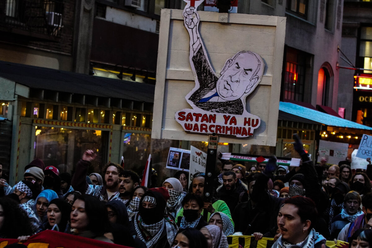 Crowd of protesters wearing keffiyehs march through street with a large cardboard sign of Israel's prime minister Benjamin Netanyahu above read letters that read “Satan Ya Hu War Criminal”