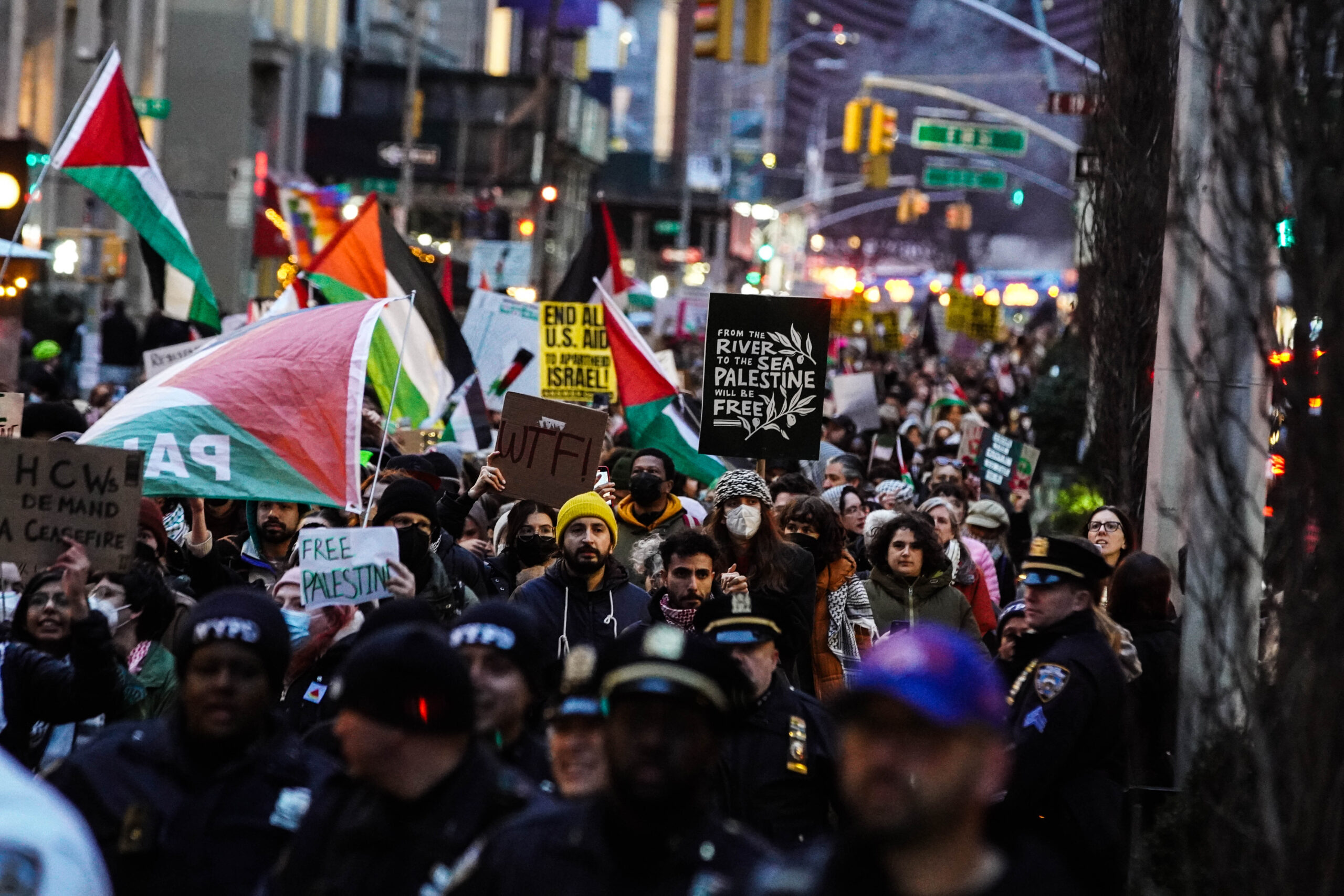 Large crowd of the protesters marching through the streets with NYPD at the the front many flags and and signs are raised in the air a prominent one reading “from the river to the sea Palestine will be free”