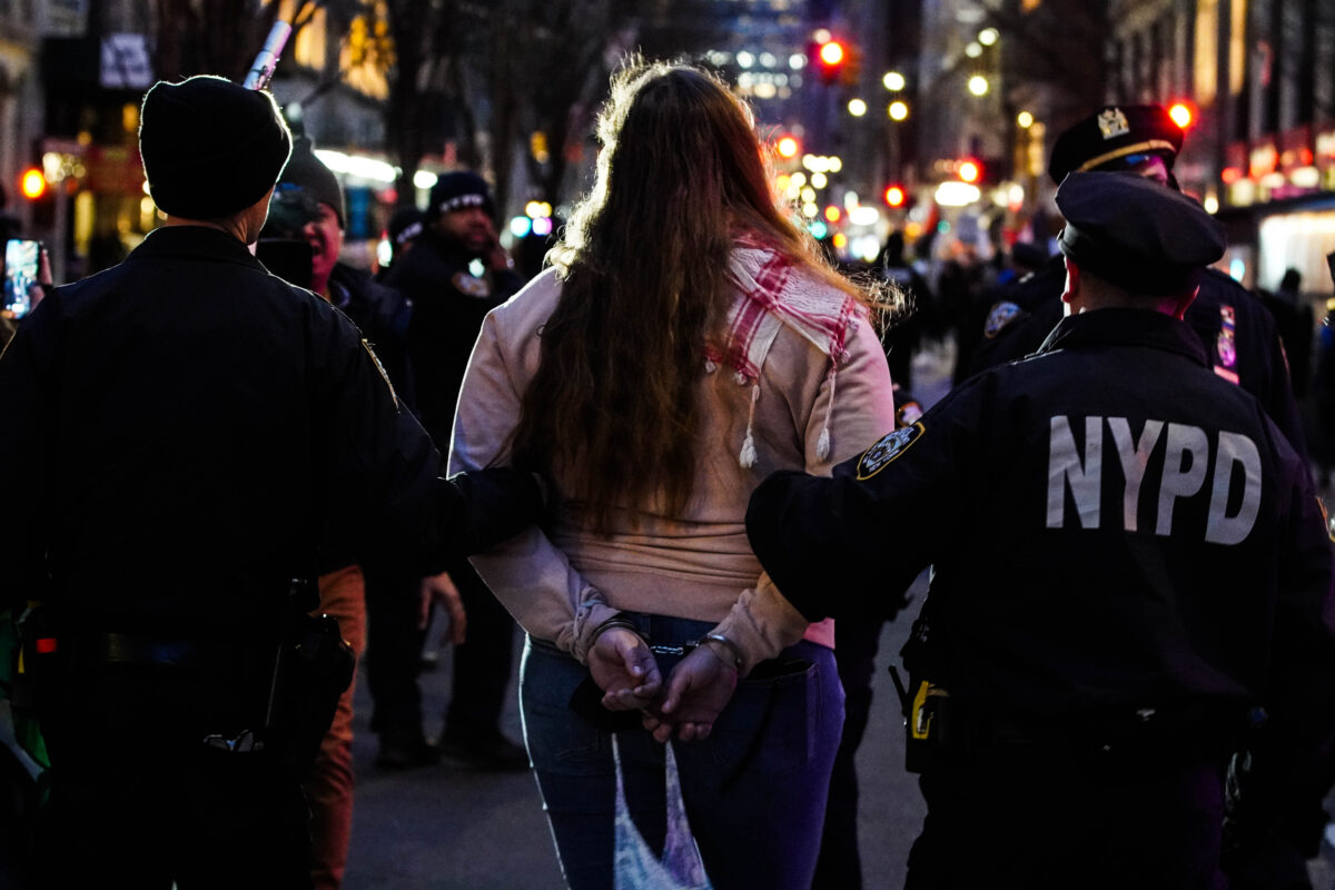 A protester with their hands cuffed behind their back being escorted away from the crowd by two NYPD officers 