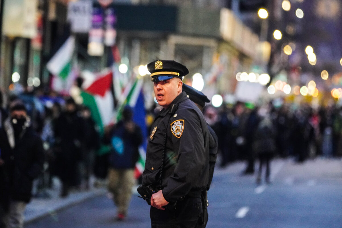 NYPD officer looks back over his shoulder into the camera with a blurred crowd of protesters in the background 