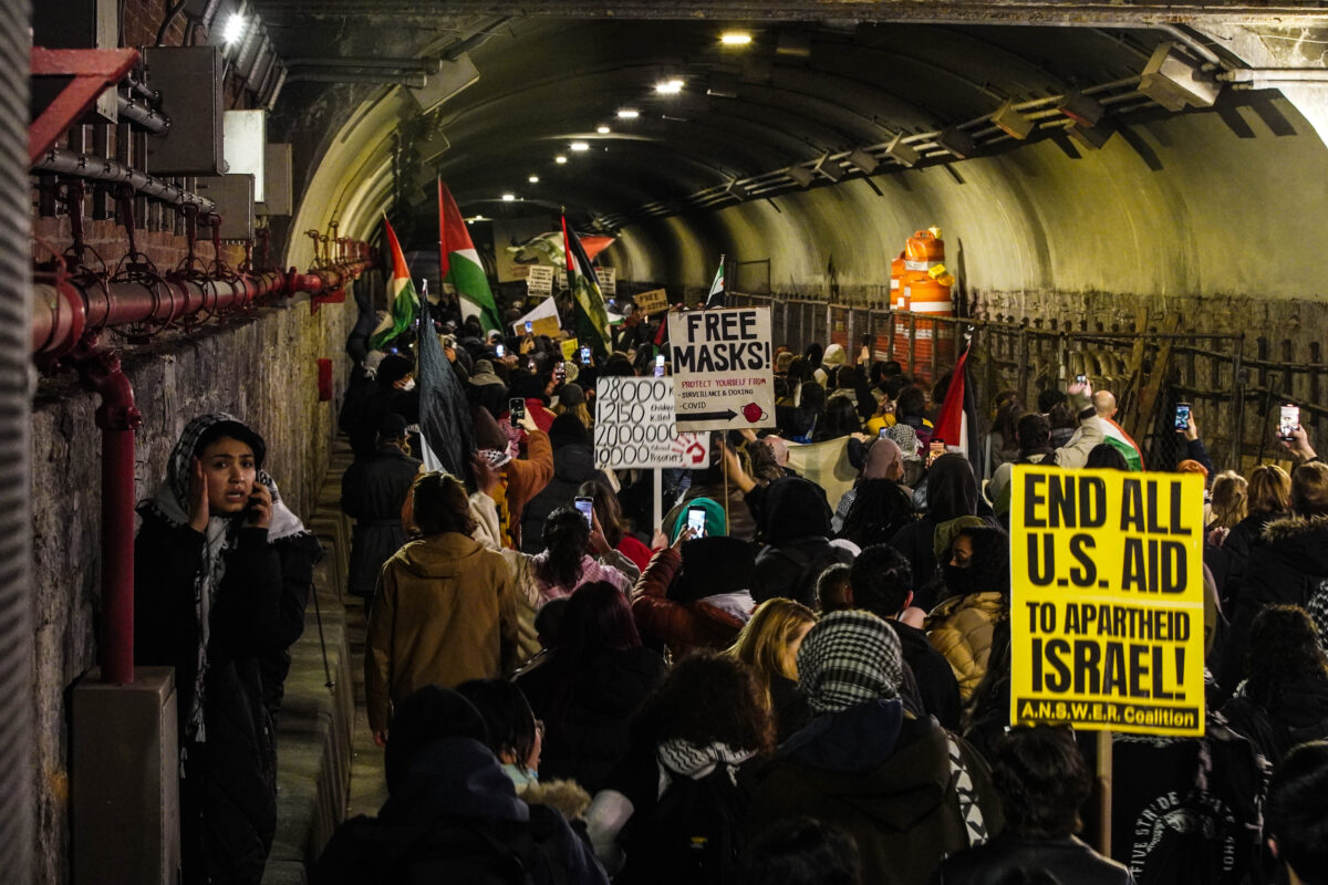 Crowd of protesters flows through a dimly lit tunnel, a young girl wearing a keffiyeh stands off to the left, holding a phone to her ear attempting to locate a friend